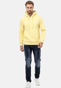 Gelbe Kapuzenjacke mit Kängurutasche, Zugbändern und gerippten Bündchen, kombiniert mit dunkelblauen Slim-Fit-Jeans und weißen Sneakers.