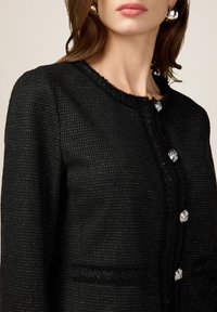 Chaqueta negra texturizada con escote redondo, adornada con botones plateados. Presenta bordes deshilachados y un ajuste estructurado.