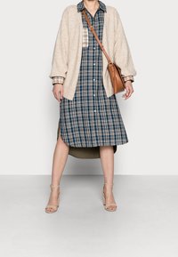 Un cardigan en tricot beige sur une robe chemise à carreaux bleu marine avec des accents blancs et beige, associé à des sandales à talons beiges et un sac bandoulière marron.