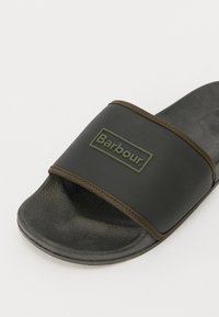 Czarne klapki z grubym, zielonym paskiem z gumy, na którym znajduje się logo Barbour, wyprofilowaną wkładką oraz gładką czarną podeszwą.