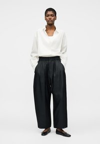 Sieviete valkā vaļēju baltu pogājamu kreklu, melnas plata pieguļoša stila bikses un melnas dzīvokļveida kurpes, stāvot pret vienkāršu gaišu fonu.