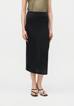 Femme portant une longue jupe noire, un top sans manches vert olive et des sandales à talons noires avec des détails en chaîne dorée, debout devant un fond blanc.