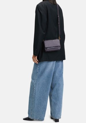 Personne portant un blazer noir, un jean bleu à jambes larges, des chaussures noires, et portant un petit sac bandoulière violet texturé.