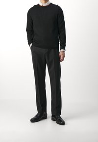 Homme portant un pull noir par-dessus une chemise blanche à col, un pantalon noir et des chaussures habillées noires, debout devant un fond blanc uni.