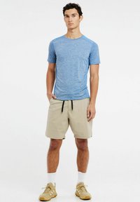 Modrá športová košeľa s krátkym rukávom vyrobená z melírovaného materiálu, v kombinácii s khaki šortkami. Model stojí s rukami v vreckách, obutý v béžových teniskách.