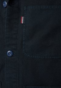 Tissu de chemise bleu marine foncé avec boutons visibles, une poche frontale, et une petite étiquette rouge et blanche portant l'inscription "SERGE BLANCO".