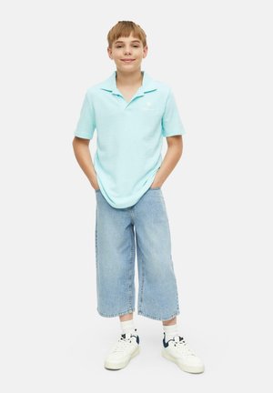 Junge steht mit den Händen in den Taschen, trägt ein hellblaues Polohemd, weite, gekürzte Jeans, weiße Socken und weiße Sneakers.