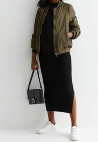 Kvinna i olivgrön bomberjacka, svart ribbad midikjol med sidoklyfta, vita sneakers och bärande på en svart handväska.