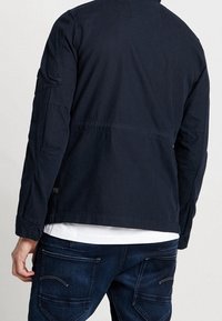 Mörkblå jacka med avslappnad passform, med hög krage, axelpartier och en slät textur. I kombination med mörkblå jeans.