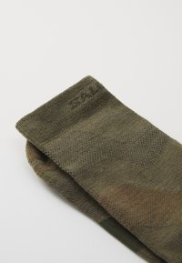 Karo dėtose kojinėse įvairių atspalvių žaliai ir rudos spalvos, pagamintose iš tekstūruotos mezginio medžiagos, su sulankstytu kraštu, ant kurio matoma prekės ženklo etiketė.