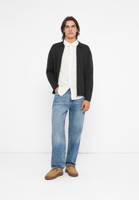 Cardigan cinza escuro sobre uma camisa branca de colarinho, combinado com jeans de perna larga azul clara e sapatos slip-on bege com solas texturizadas.