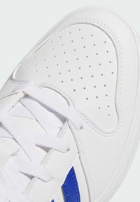 Bijela tenisica s perforacijama na prstu i bijelim vezicama, s plavim detaljem pruge sa strane.