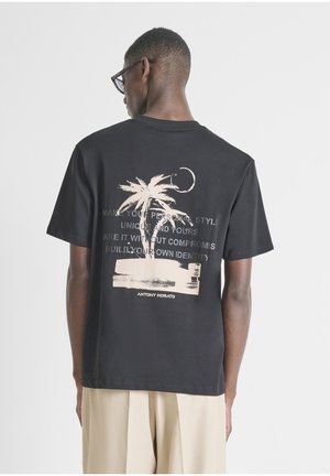 Hombre con camiseta negra con diseño de palmera beige y luna, texto, y pantalones beige, de espaldas frente a un fondo claro y sencillo.