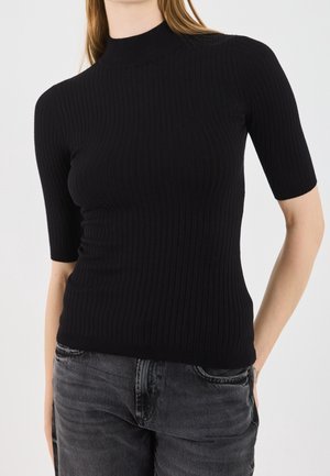 Femme portant un top noir côtelé moulant à manches courtes et un jean noir délavé, debout avec une main derrière le dos.
