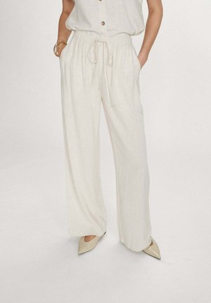 Femme portant un pantalon ample en lin beige clair avec une taille à cordon, associé à des escarpins pointus beiges et des bracelets dorés à un poignet.