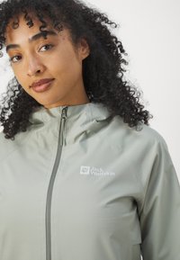 Lichtgroene jas met een rits aan de voorkant, capuchon en logo op de linkerborst. Gladde textuur met naden en een getailleerd ontwerp.