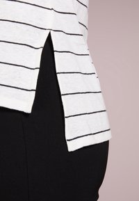 Weißes Hemd mit dünnen schwarzen horizontalen Streifen, in schwarze Hose gesteckt, zeigt ein seitliches Schlitzdetail am Hemdenrand vor einem schlichten Hintergrund.