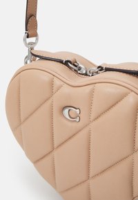 Srdcovitá taška z světle béžové kůže s prošívaným vzorem, stříbrným kováním a hnědým akcentem na popruhu. Zapínání na zip.