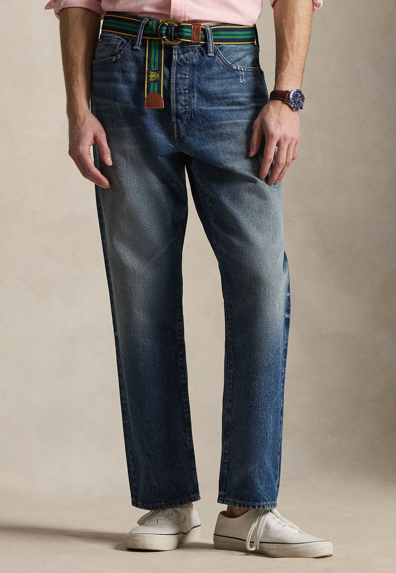 Άντρας που φοράει ξεβαμμένο μπλε τ ζιν με πράσινη ριγέ ζώνη, λευκά αθλητικά παπούτσια, ροζ πουκάμισο και ρολόι χειρός σε απλό φόντο.