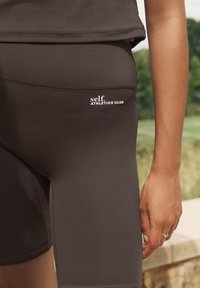 Fekete atlétikai rövidnadrágok, testhezálló kialakítással és sima textúrával, amelynek derékpántján fehér színű "self. ATHLETICS CLUB" logó található.