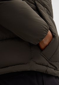 Giacca puffa verde scuro con una texture liscia e una tasca laterale. Cuciture visibili e un'apertura nascosta nella tasca.