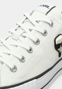 Witte vetersneakers met zwarte accenten, metalen oogjes en een cartoonlogo van een persoon met een zonnebril op de zijkant.