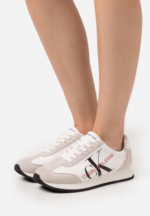 Zapatillas blancas con detalles en ante beige, con un destacado logo "CALVIN KLEIN JEANS" en negro y rojo, diseño clásico con cordones, suela negra.