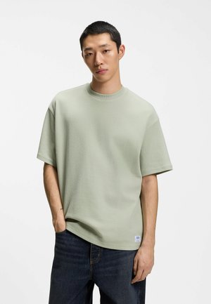 Young man with short hair wearing a light green textured short-sleeve shirt and dark jeans, standing with one hand in pocket.