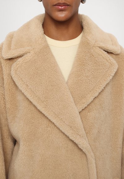 Beige faux fur coat with a wide lapel, soft texture, and double-breasted design. Styled over a light yellow top, with no visible fastenings.
