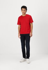 T-shirt en coton rouge à manches courtes et poche poitrine, associé à un jean en denim foncé et des baskets blanches, se tenant devant un fond uni.