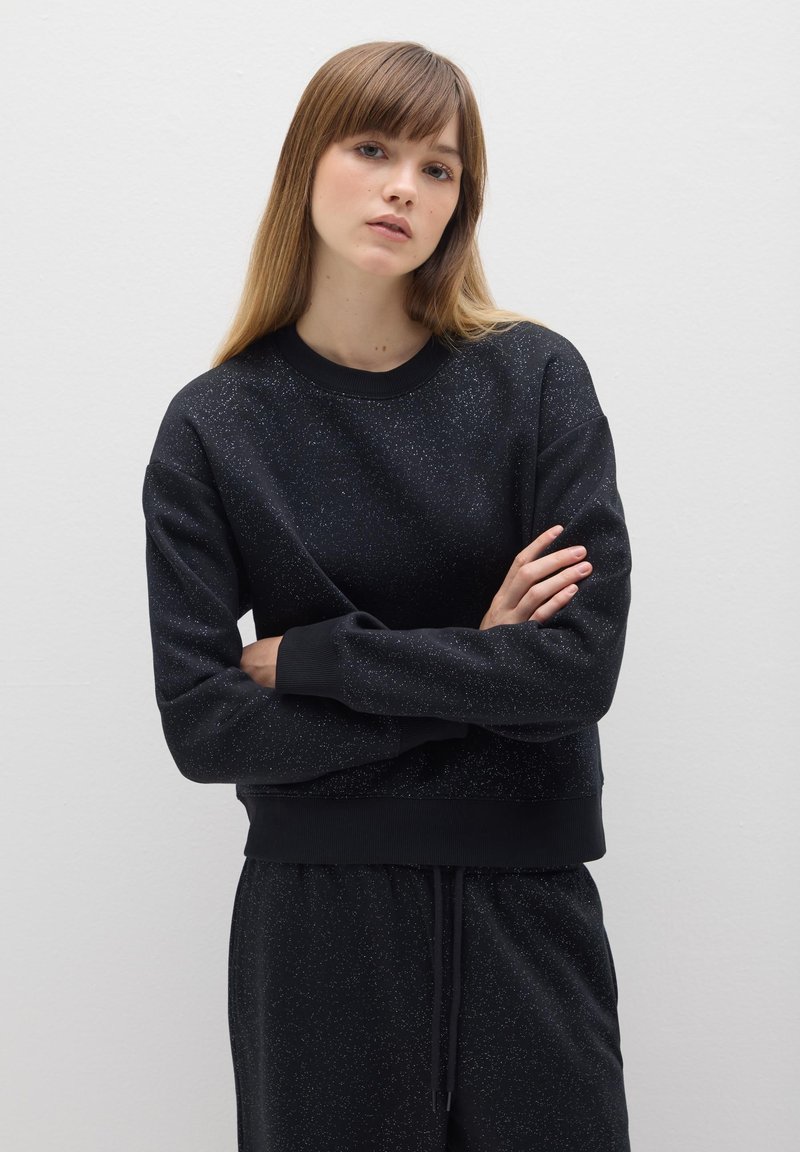 Sweatshirt noir avec une texture tachetée, un col ribber et des poignets. Associé à un pantalon assorti, créant un look cohérent.