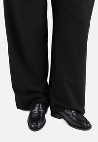 Mocasines de cuero negro con un acabado texturizado y un detalle plateado, usados con pantalones anchos negros que muestran un ajuste relajado.