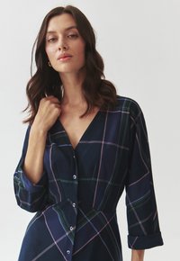 Robe écossaise bleu marine avec décolleté en V, manches longues et fermetures à bouton. Présente des lignes vertes, roses et bleu clair dans un motif à carreaux.