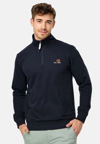 Sudadera de forro polar azul marino con cremallera de un cuarto, que presenta un pequeño logo bordado en naranja y una textura suave. Corte casual con puños acanalados.
