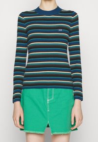 Maglione a righe coste nei toni del verde petrolio, blu navy e marrone con collo a girocollo blu a contrasto. Abbinato a una gonna di denim verde.