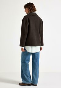 Giacca oversize marrone scuro con colletto a punta, indossata sopra una camicia a righe bianche, abbinata a jeans blu larghi e stivaletti alla caviglia.