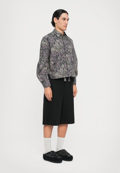 Hemd mit Knopfleiste in Dunkelgrau mit floralem Muster, kombiniert mit schwarzen knielangen Shorts und schwarzen bestickten Slippern mit weißen Socken.