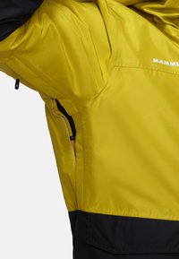 Ljust gul jacka med en svart nederdel, med en slät yta, dragkedjefickor och en logotyp på övre bröstområdet.