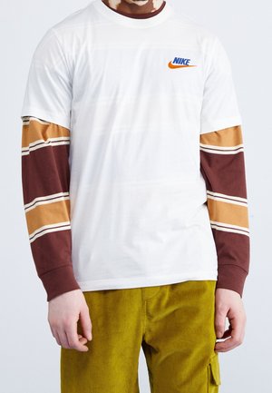 Hvid Nike t-shirt med kort ærme og orange logo, lagdelt over lange brune og beige stribede ærmer, parret med sennepsfarvede fløjl bukser.