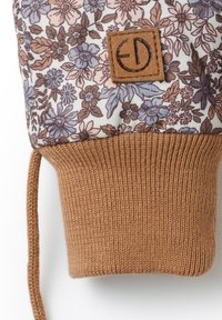 Mönstrad textil i beige med blommönster i brunt och blått, med en tan stickad manschett och en fyrkantig läderlapp med logotyp.