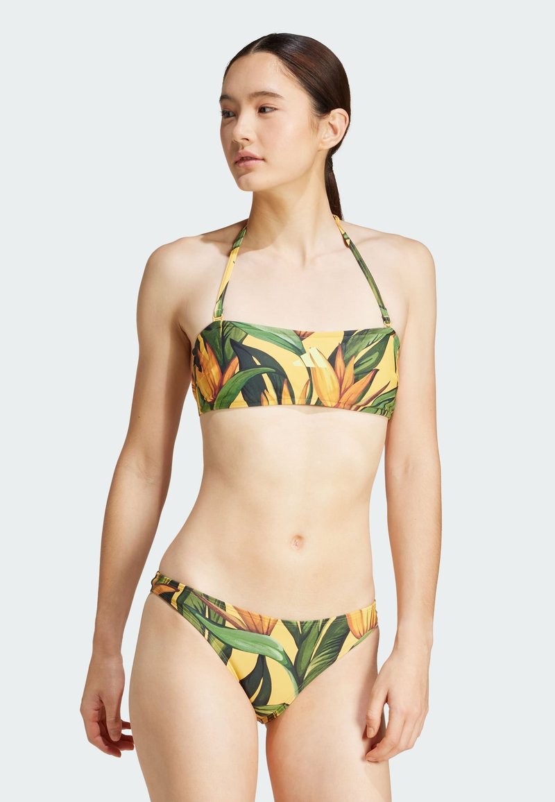 Bikini set s žlutým základem a vzory tropických listů a květin. Vrchní část má bandeau design s nastavitelnými ramínky; spodní část je s nízkým pasem.