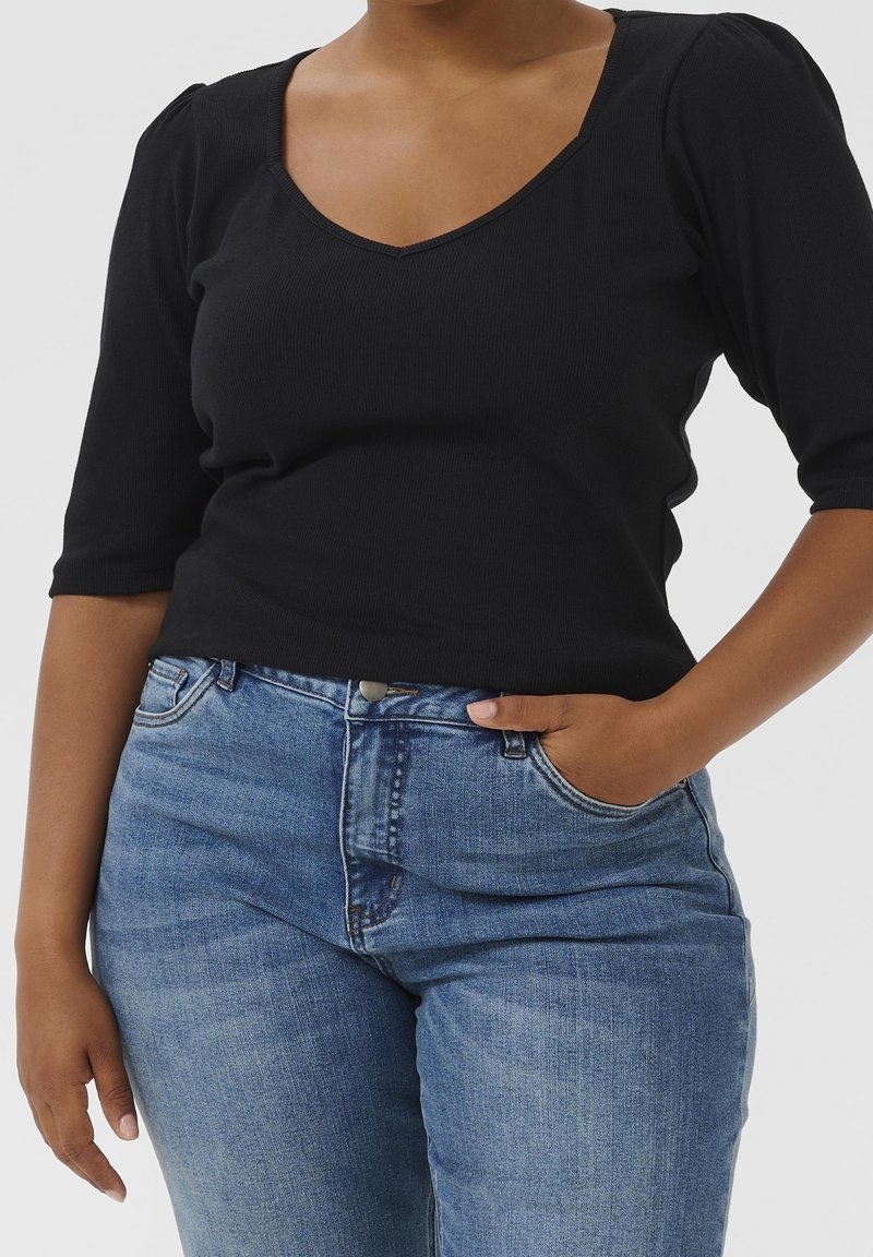 Femme portant un haut noir côtelé à col en V avec des manches aux coudes et un jean bleu taille haute, une main dans la poche.