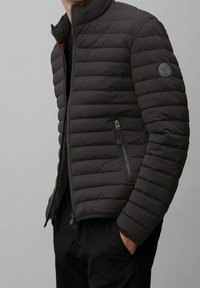 Giacca a vento nera imbottita con colletto alto, dotata di chiusura a zip frontale, tasche laterali con zip e una patch con logo sul braccio sinistro.