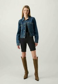 Ung kvinne står, iført blå denimjakke, sorte shorts og brune knelang støvler, mot en enkel lys bakgrunn.