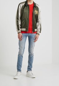 Chaqueta bomber de satén verde con acentos crema y bordado de tigre, combinada con una camiseta roja, jeans de mezclilla azul claro y zapatillas blancas.