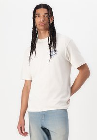 T-shirt beige à manches courtes avec le texte "Chasing Dreams" en bleu, coupe décontractée et en coton doux. Assorti à un jean bleu clair.