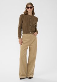 Ženska oblečena v rjav volneni cardigan, bež široke kordne hlače, rjave koničaste čevlje in temne sončne očala, stoji z eno roko v žepu.