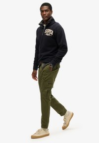 Marineblå fleece zip-sweater med "SUPER DRY" broderi, grønne cargo-bukser og beige sneakers med tekstureret design.