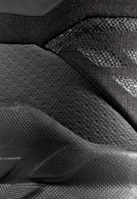 Svart sportskos övre del med texturerade tygpaneler, släta syntetiska sektioner och upphöjda mönster för ökat grepp och designdetaljer.