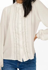 Weißes Blusenoberteil mit hohem Kragen, vertikalen Falten, Knopfverschluss, Spitzen-Einsatz und puffärmeligen Ärmeln, kombiniert mit blauen Jeans.